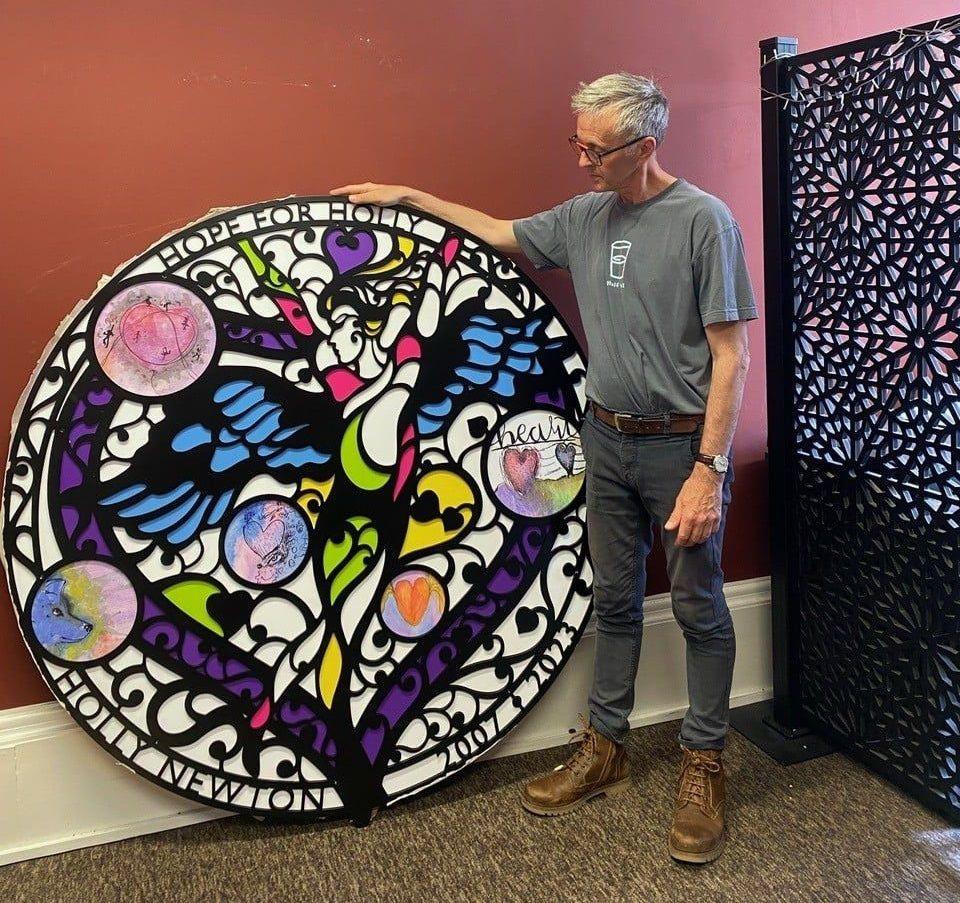 Artist Steve Pardue standing with the large circle piece. It shows a dancer with wings with various colourful designs surrounding it. At the top it reads 'Hope for Holly' and at the bottom. The design is largely black and white with bits of pink, green, yellow, blue and purple.