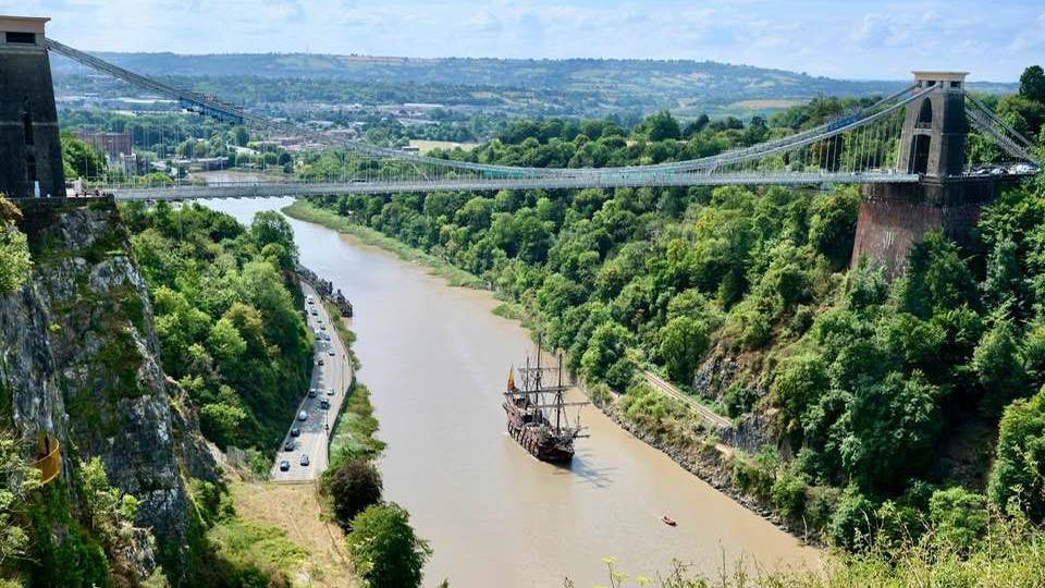 A Spanish galleon ship is pictured from above as it sails under the Clifton Suspension Bridge down the River Avon heading for the Severn Estuary. The river is at high tide and the water is a grey/brown colour