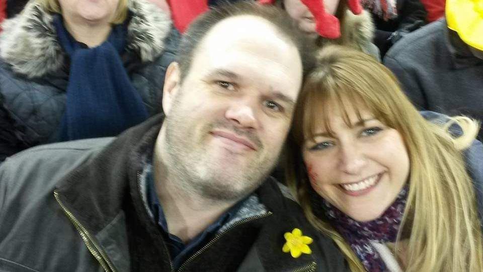 Sam Morgan has short brown hair and dark eyes, Karen Morgan has dark blonde hair, a fringe and blue eyes. Sam is wearing a yellow daffodil on his dark coat, and Karen has a Welsh dragon painted on her cheek and is wearing a purple scarf