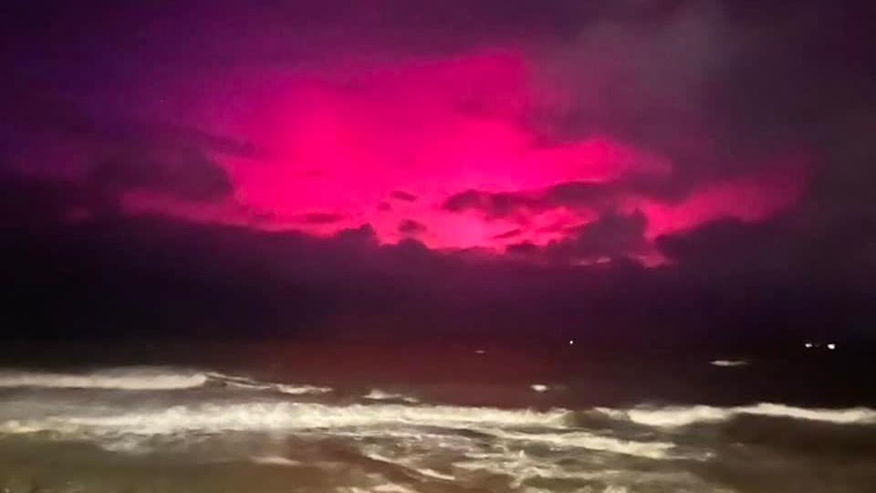 The aurora borealis shining over the sea in Westward Ho!. The aurora has a pink-red hue. Waves are crashing on the beach in the foreground.