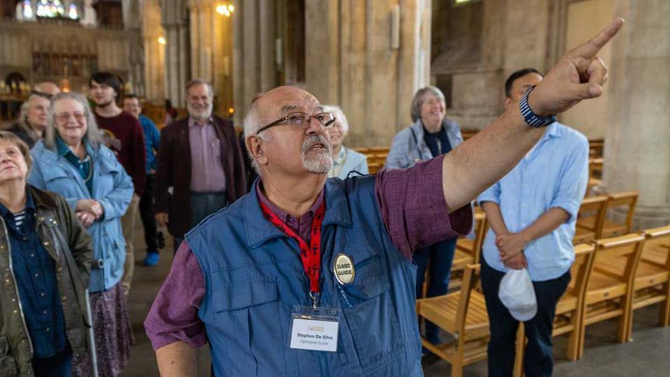 Stephen de Silva wears a maroon shirt and a blue gilet on which is attached gold coloured badge with the word guide on it. He also has a red lanyard round his neck attached to a plastic case containing his name badge.  He has grey hair with a short grey moustache and beard and is balding. He is wearing clear-rimed glasses and is pointing out something to visitors inside the cathedral.