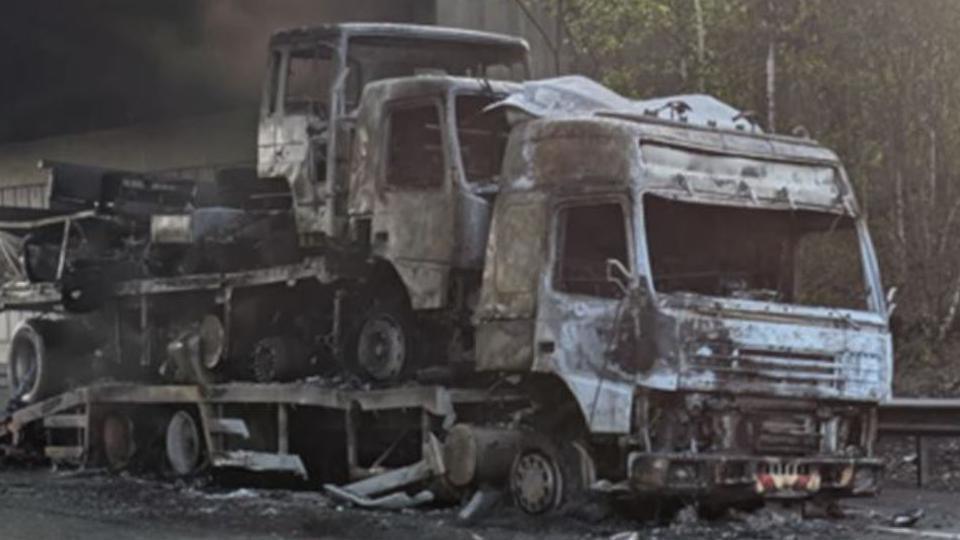 Charred shell of lorry with a further two being carried on its trailer. The lorry is at the side of a road under a bridge