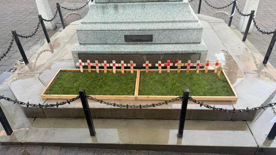 The remedied work at the war memorial. It shows two neatly trimmed rectangles of turf placed inside two wooden boxes, with several wooden crosses baring poppies stuck into the grass.