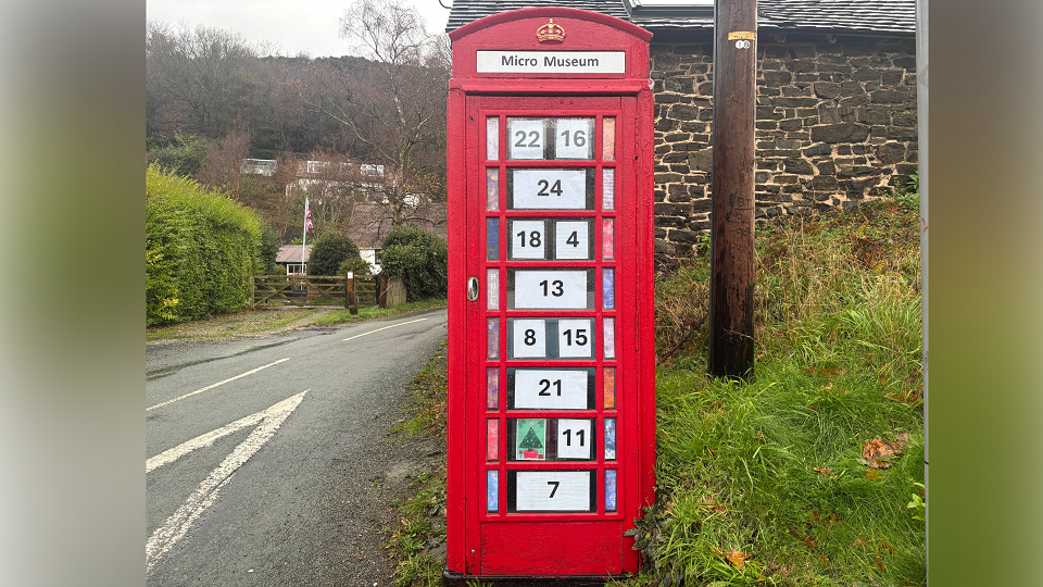 A red phone box on a grass verge next to a country road. In each window is a piece of paper with a black number on it. In place of one number, there is a piece of artwork depicting a Christmas tree