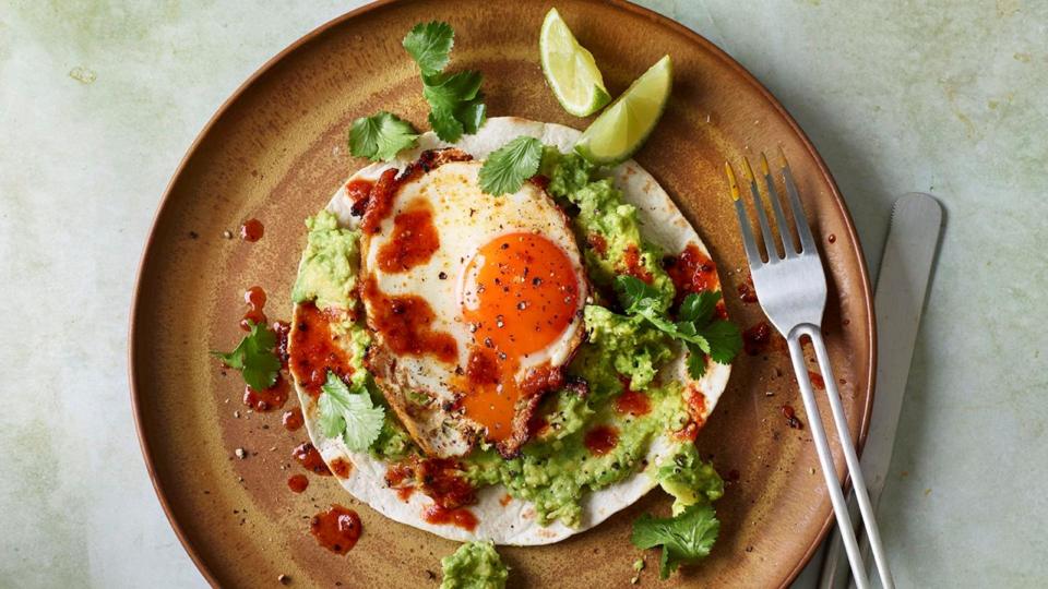 A fried egg, coriander leaves and smashed avocado on a tortilla, all sitting on a brown plate