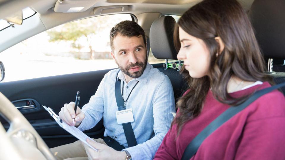 A young woman sitting behind the wheel of a car about the take her driving test. A male driving test examiner is going through a checklist first.