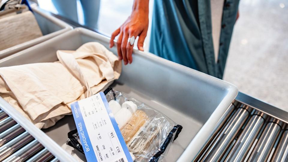 Stock photo shows a person's hand moving along a grey tray containing personal items and a boarding pass including a clear plastic bag containing liquids, on a roller in an airport security zone.