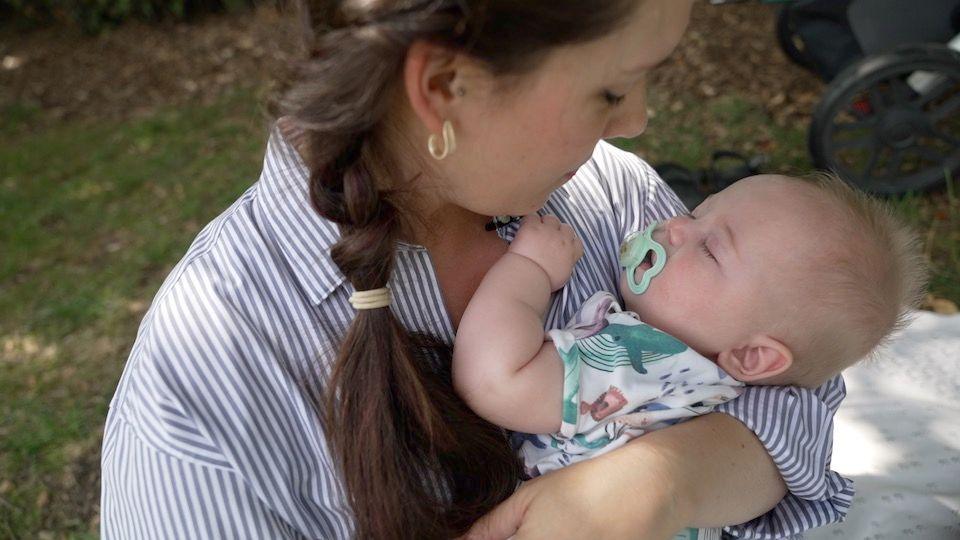 A mother in a blue and white striped shirt, dark hair braided in a plait, cradles her sleeping baby who is wearing a onsie with sea animals on it. He is sucking a dummy.