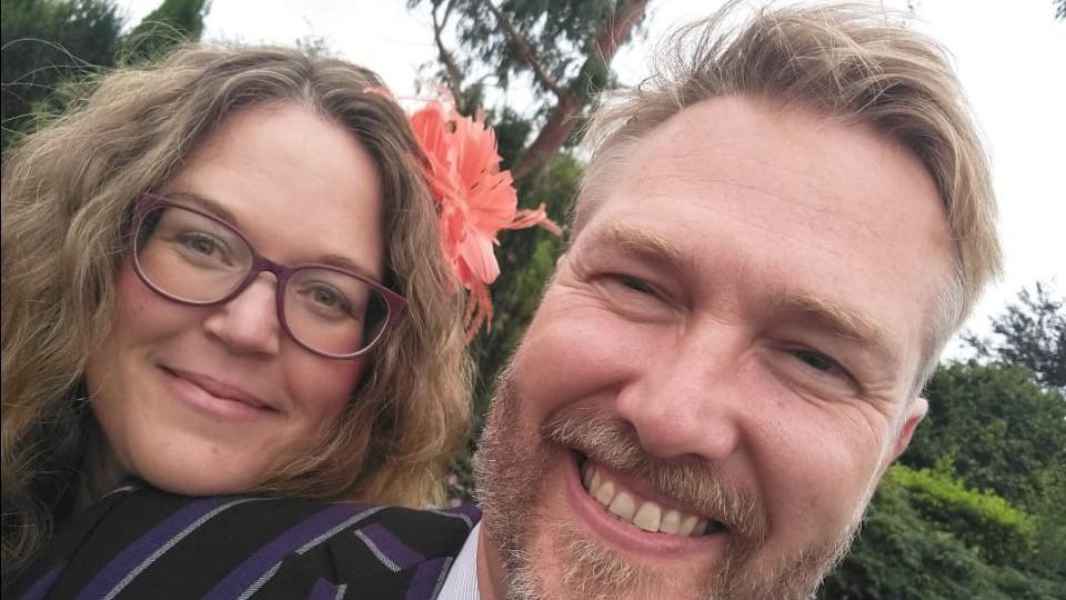 A close-up image of Shiela Lorking who is resting her head on her husband's shoulder. She has shoulder length brown hair with a peach-coloured flower pinned to her hair and is smiling. Her husband has short blonde hair with a blonde beard. Trees and bushes are seen behind them.