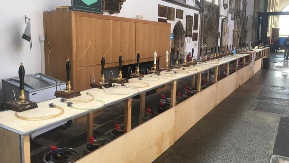 A long line of more than 20 beer pumps attached to a long wooden temporary bar inside a church. Old stonework and windows can be see in the background.