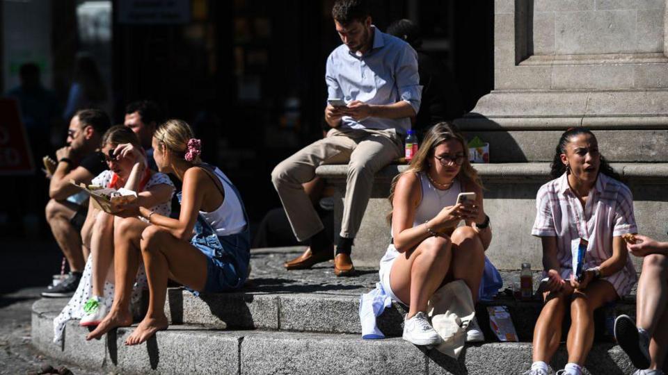 Men and women sit on the steps of a fountain in the sunshine while eating their lunch and checking their phones. A number of women are wearing shorts.