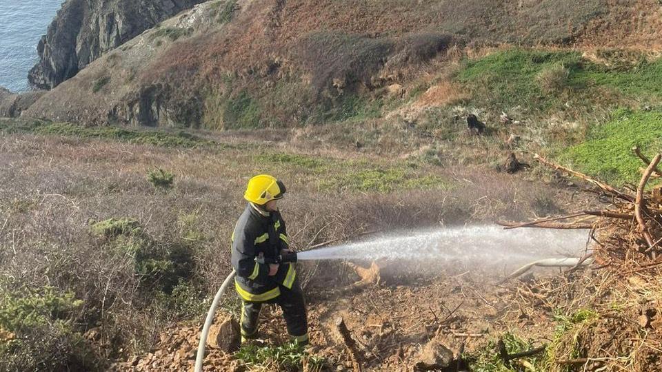A fire fighter is holding a hose pipe and spraying water on the grass area on the cliff side. The fire fighter is wearing protective clothing and a yellow hard hat.