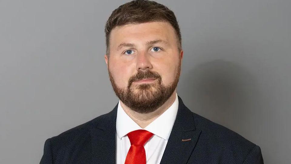 Blackpool South Labour MP Chris Webb  in his UK Parliament portrait. He is standing in front of a plain grey background wearing a dark blue suit, white shirt and red tie. He has short brown hair and a shot brown beard