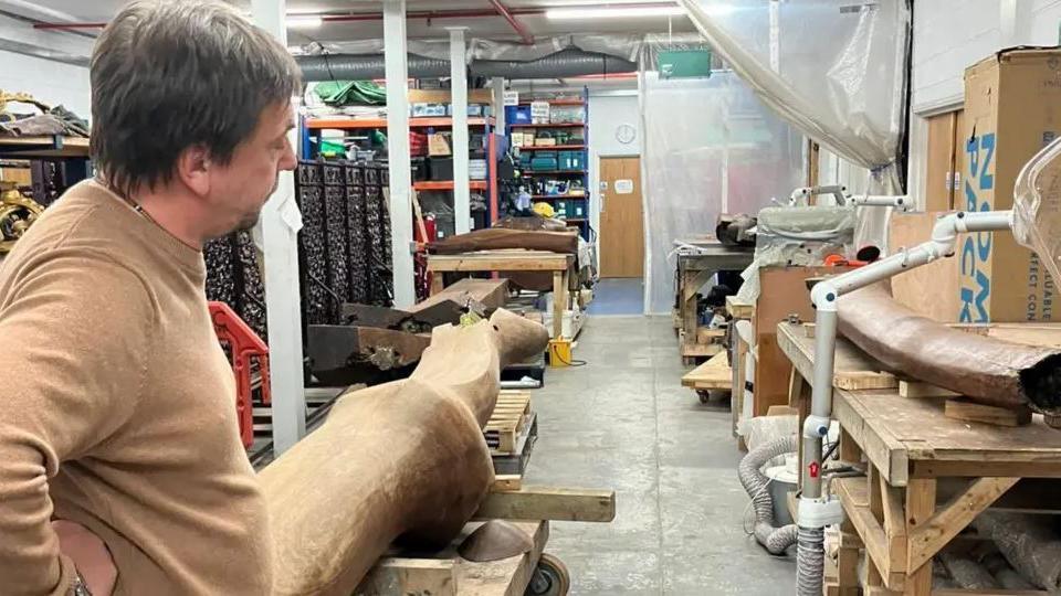 A man wearing a light brown sweater, standing by a wooden sculpture that looks like a log, inside a workshop.