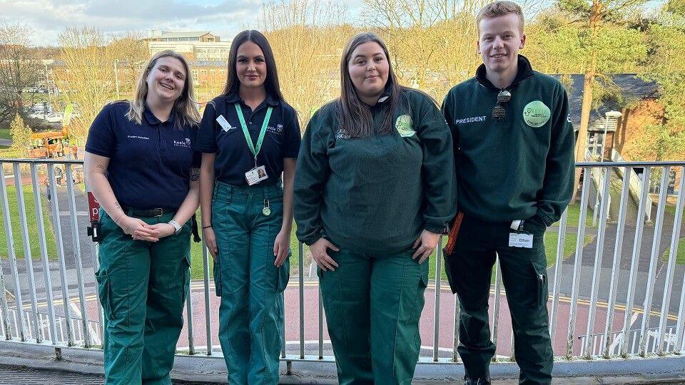 Three women and one man dressed in green paramedic outfits, standing in front of a fence above a road. 