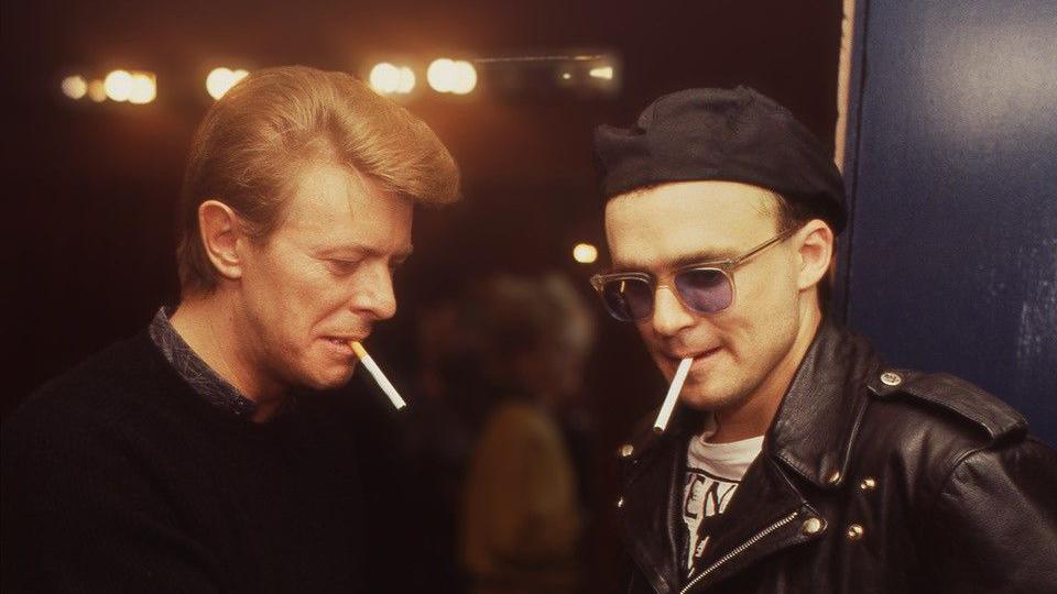 David Bowie and a man with a black hat and blue glasses. They both have cigarettes in their mouths