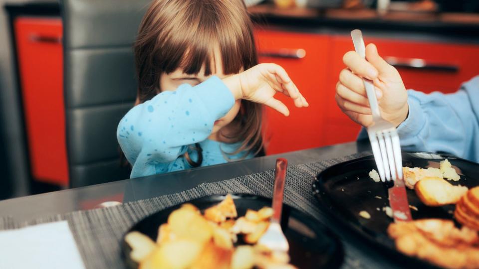 A small child sitting in front of a plate of food, covering her face with her arm.