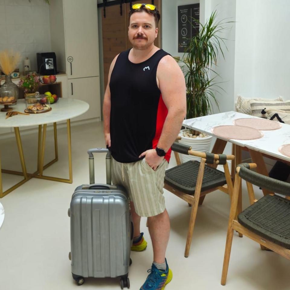 Hywel Roberts, wearing vest and shorts and with sunglasses on his head, with a suitcase, in a room