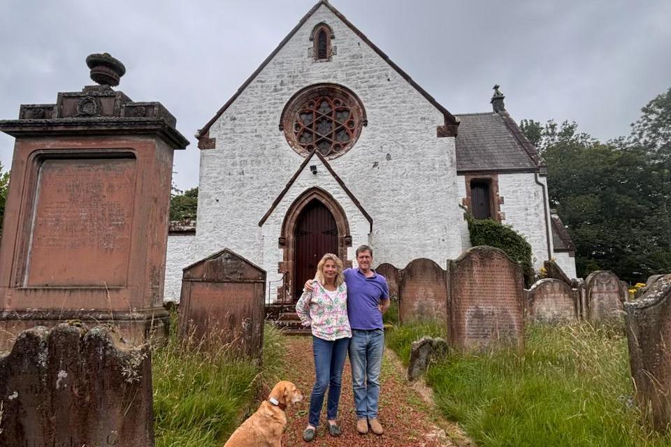 Kirsteen Mitchell and her husband Andrew standing in front of a whitewashed church surrounded by gravestone with a golden-coloured dog by their sides