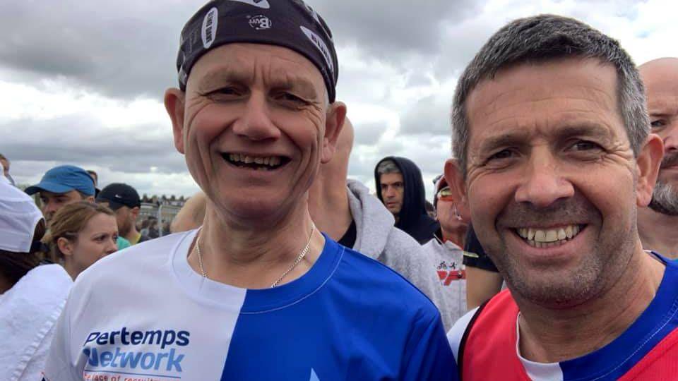 Two men in blue and white halved running shirts smile at the camera. One is wearing a black bandana. There are lots of people behind them.