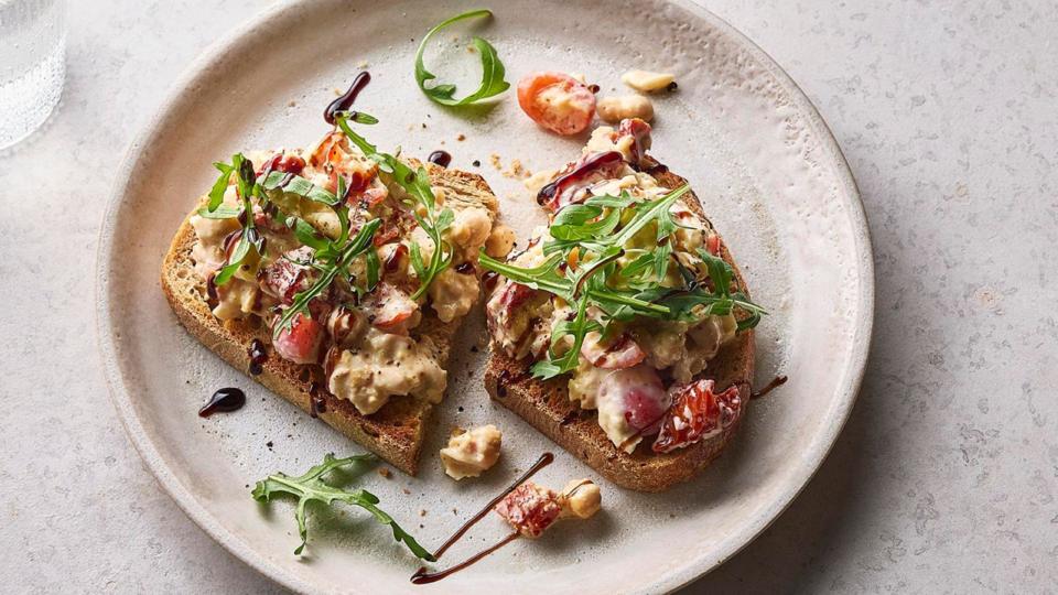 Two pieces of toast on a white plate topped with smashed chickpeas, cherry tomatoes and rocket