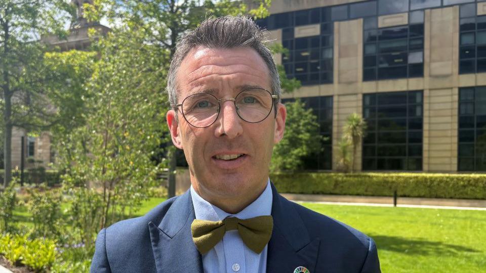 Minister Muir has short dark hair, round metal glasses and is wearing a navy suit jacket, blue shirt and green tie.