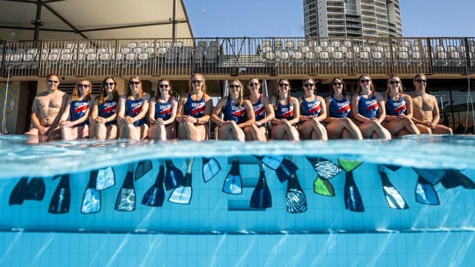 A group of people in red, white and blue swimming suits sat by the edge of an outdoor pool. The group are sat wearing flippers in the water. Half of the shot is above the water, while the other half is underwater.