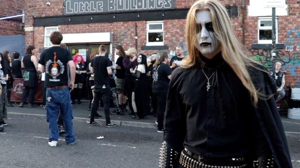 Envus is stood in front of a crowd outdoors. He is wearing all black, has long brown hair and his face painted white with black accents.