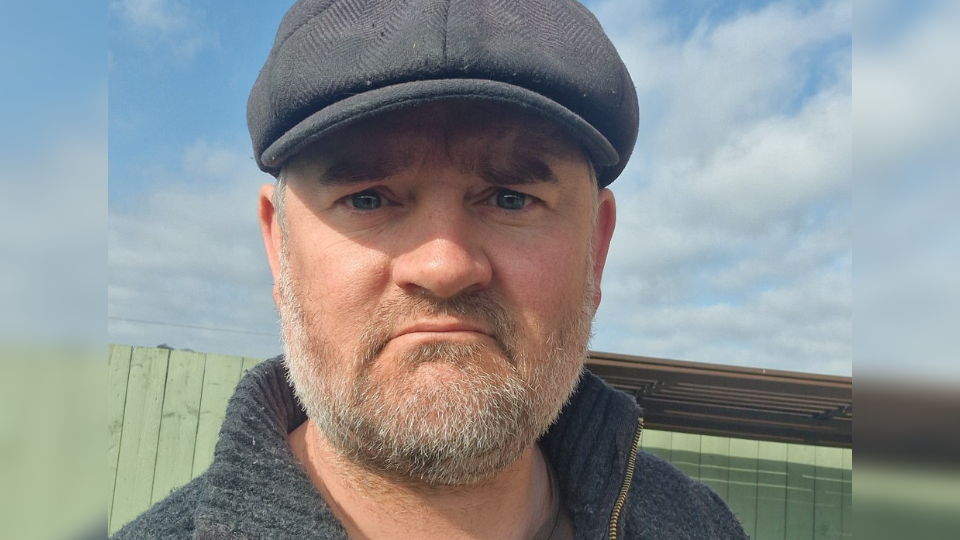 A man with a grey beard looks at the camera, with an annoyed expression on his face. He is wearing a flat cap and a grey woollen jumper. In the background is a blue sky with some clouds, and a green fence.