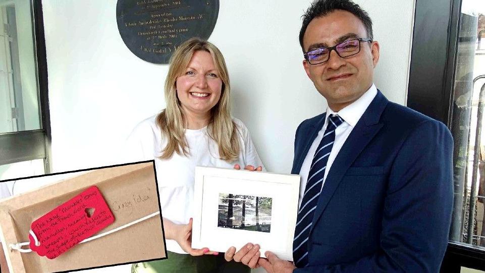 Nicola Purdie, who has medium length blonde hair and is wearing a white t-shirt, stands next to surgeon Reza Arya, who is wearing a blue suit and tie and has short dark hair and glasses. Both are smiling and are jointly holding a framed painting of a landscape showing a promenade with people, trees and the sea in the background. There is an insert into the picture of a close up of a handwritten tag on the back of the frame, next to the words "crazy idea".