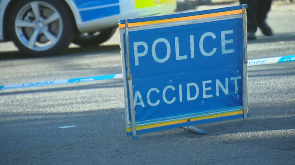 A police accident sign, which is blue with white writing, on a road in front of white and blue police tape. The wheel of a police car is visible in the background as are the feet of an officer.
