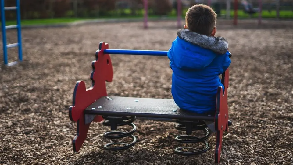 Little boy in a bright blue coat, facing away from us, sits on a spring-bound seat in a playground