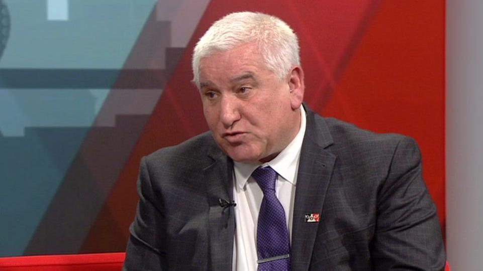 A man with short white hair and wearing a dark suit, white shirt and purple tie, is sitting in a TV studio, looking towards the presenter out of shot to the left. He has a pin on the left lapel of his jacket with a black, white then red strip on it.