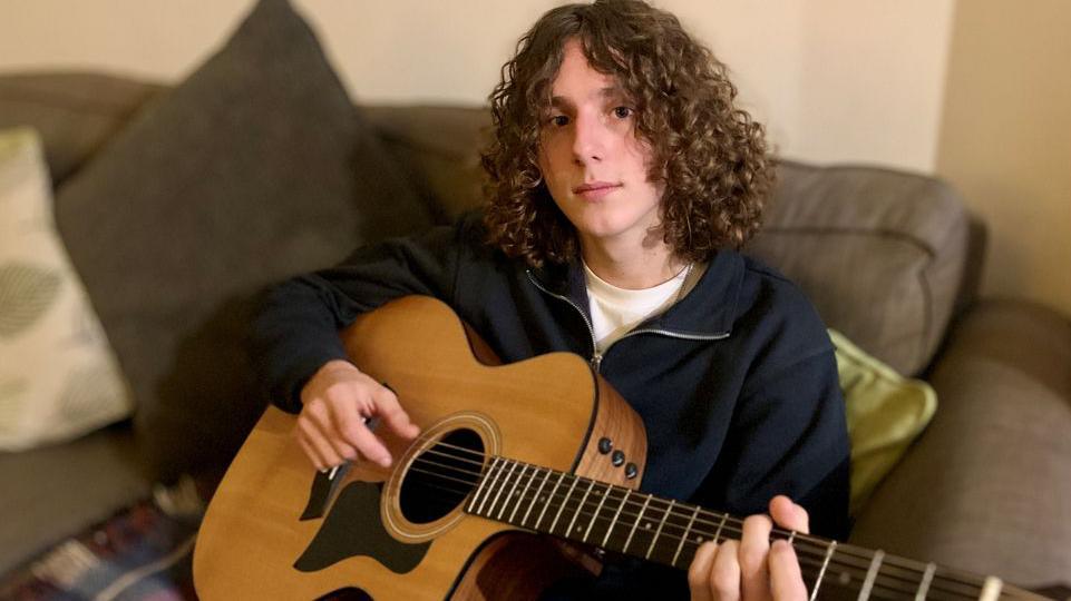 Alex Spencer with long curly hair sat holding a guitar on a sofa