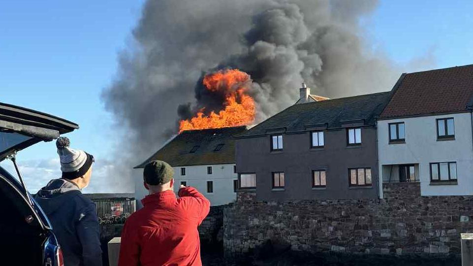 two men in blue and red jackets look at flames on the roof of buildings in the distance.