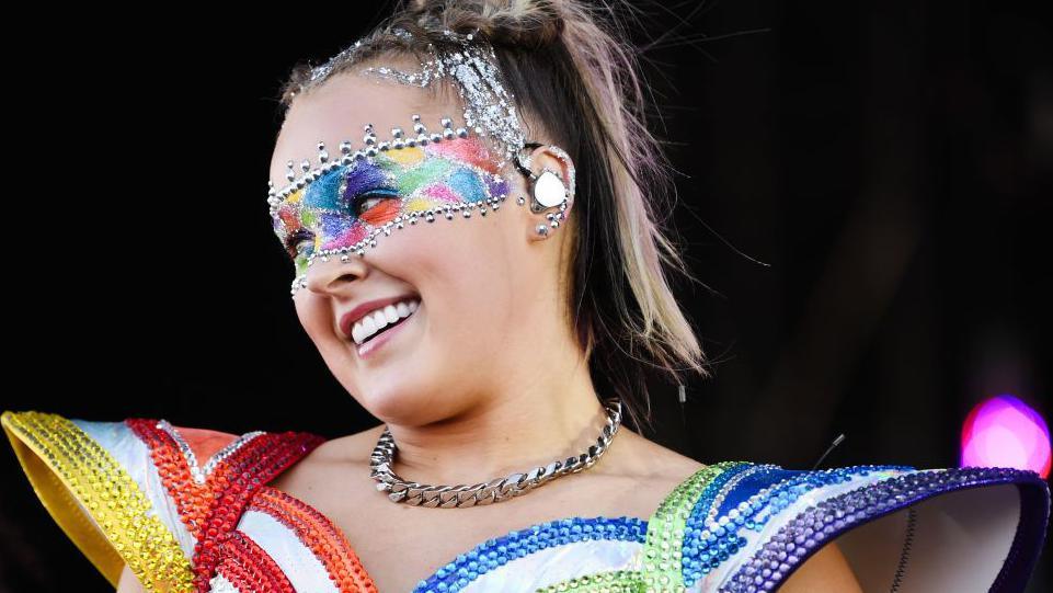 JoJo Siwa smiling while performing on stage wearing a rainbow-coloured outfit with large shoulder pads. She's wearing multi-coloured eye make-up which is framed by silver jewels and has a matching necklace around her neck.