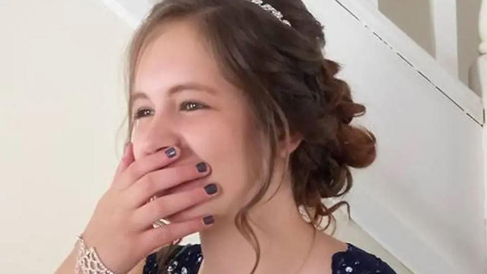Quinn Milburn-Beadle has her hand over her mouth but is smiling under the fingers. Her long dark hair is in an up do and she is wearing a bracelet.