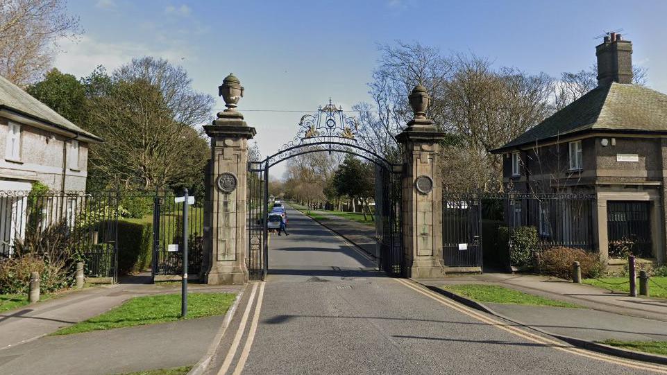 The entrance to Stanley Park, showing large iron gates between two stone columns. There are single buildings to each side and an asphalt road with double yellow lines in the middle. Some cars are parked to the left in the distance