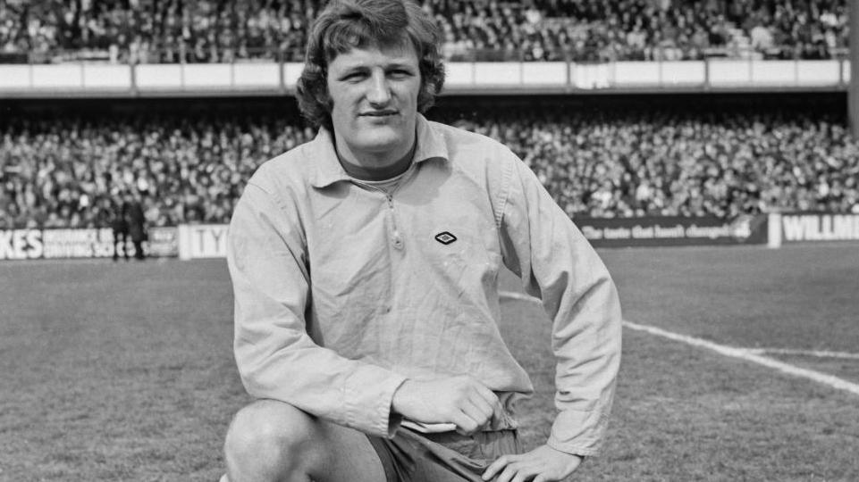 Jimmy Bone in a black and white photo before a game for Norwich City at QPR in April 1972, His left knee is on the pitch, his right knee up and his foot on the ground. His right elbow is posed on his thigh. He is wearing a long-sleeved top, and a packed terrace can be seen behind him