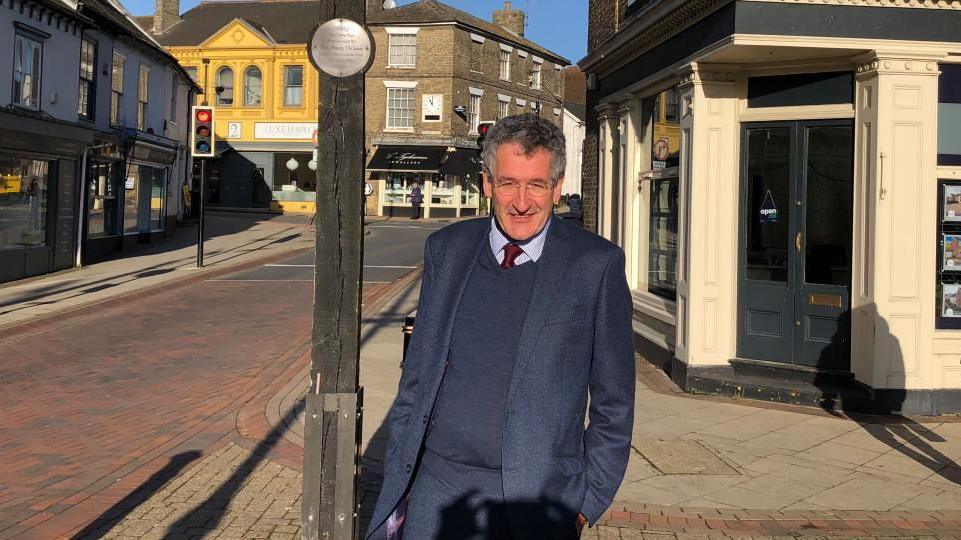 Peter Prinsley standing in the market square in Stowmarket. He is wearing a blue suit. He has grey and dark hair and is wearing glasses