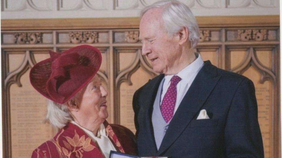 Mary Laing dressed in a red blazer with gold leaves, a white top, and a red fascinator looks up at David Laing, dressed in a navy blazer a grey waistcoat, white shirt and maroon tie in a church