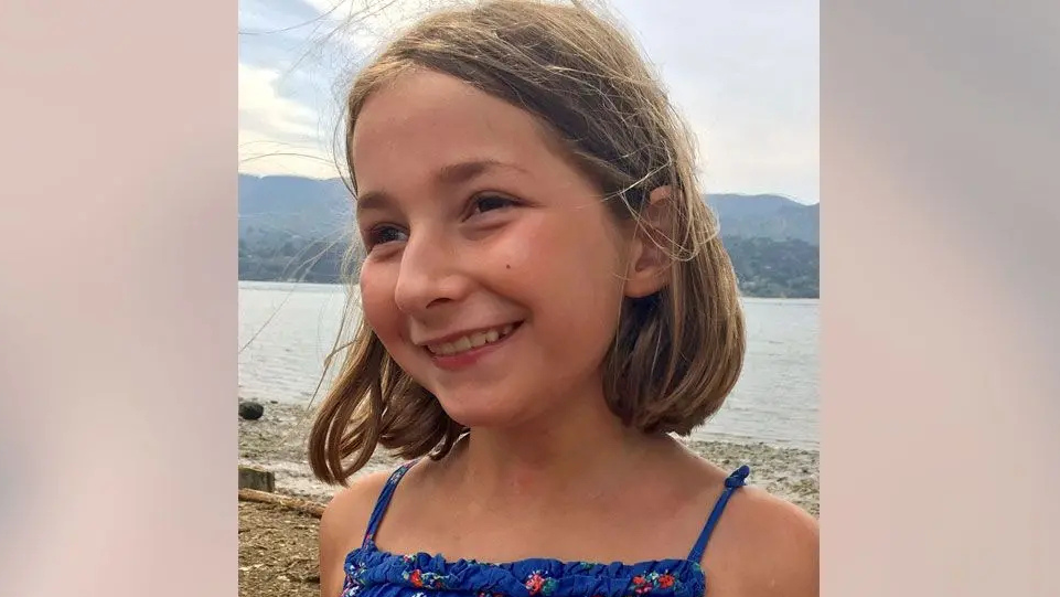 An image of a girl smiling taken in front of a lake. She has mid-brown hair in a bobbed style, she is wearing a blue floral top.