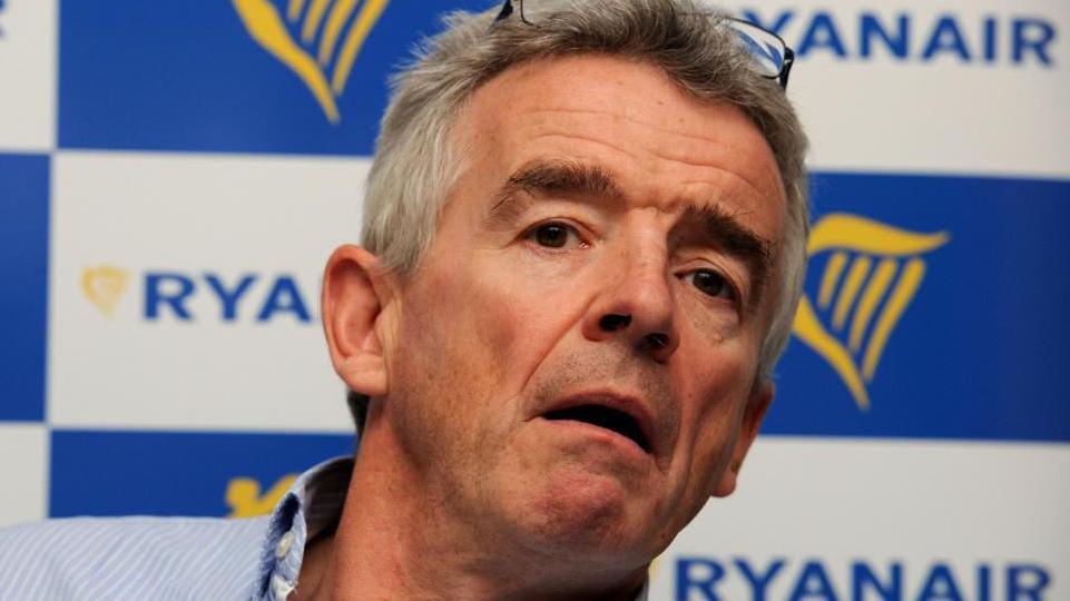 A grey-haired Ryanair boss Michael O'Leary, with his mouth slightly opened, sits in front of his company's branding. He is wearing a blue and white pinstriped shirt