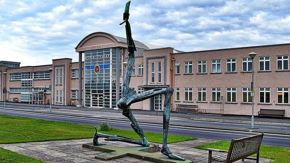 The exterior of the airport, a statue of the three legs of Man in front of the airport building, which is a block-like peach coloured building.