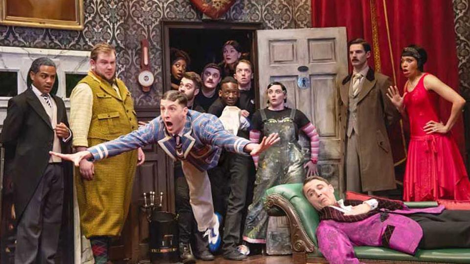 An actor in The Play That Goes Wrong falls through the air as if he has tripped up while other cast and crew look on in shock. Another actor in a pink smoking jacket lies on a chaise longue looking worried