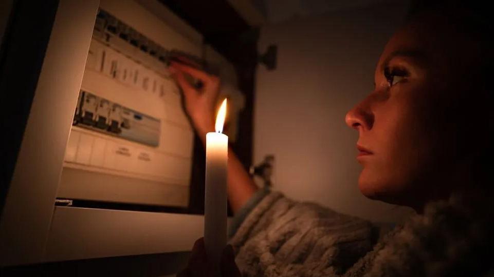 A woman in a dark room looks at a fusebox by candlelight