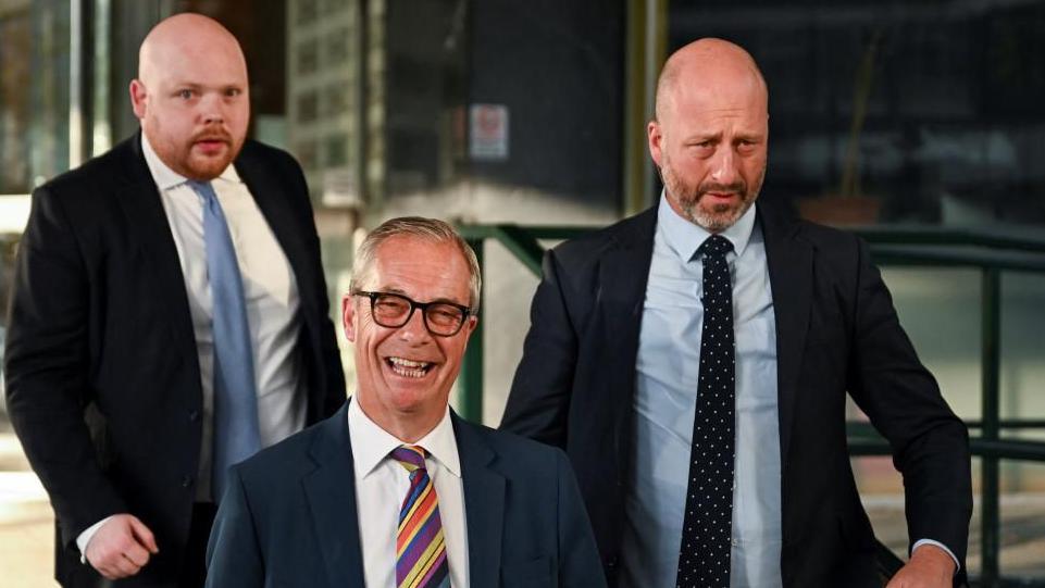 Lideri i Reform UK të Britanisë, Nigel Farage, me syze, kostum dhe kravatë me vija, i ndjekur nga dy burra me kostume, buzëqesh ndërsa largohet nga Kulla Millbank.