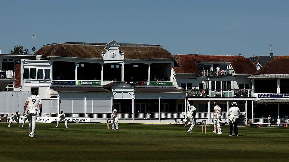 The Spitfire Ground with cricketers playing dressed in all white, with blue sky