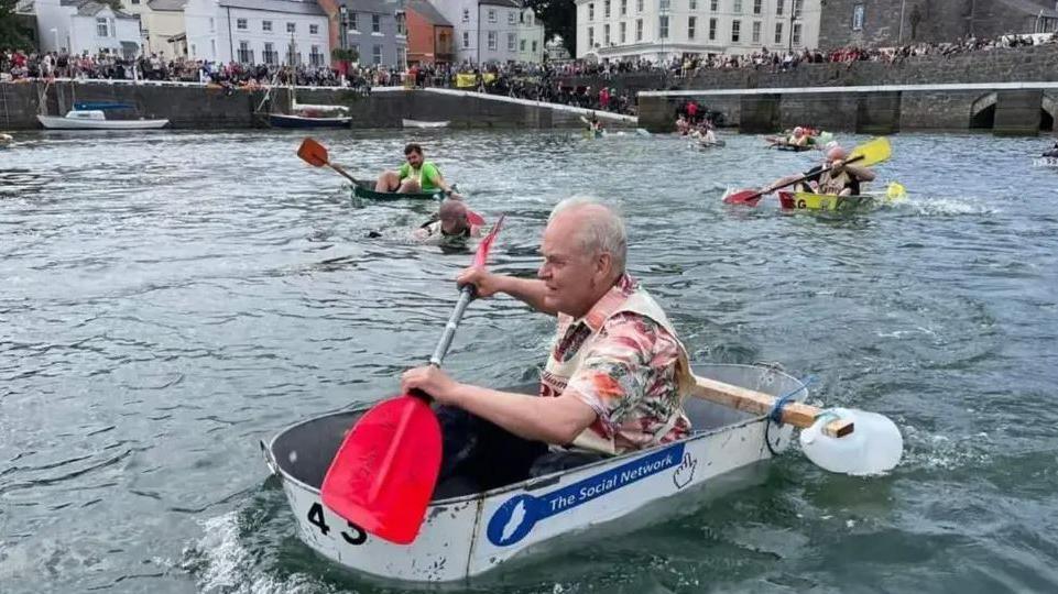 A man in a tin bath paddles in Castletown Harbour, there are other competitors in the race behind him in the water, with one man swimming after his bath has sunk. The harbour walls are lined with hundreds of spectators.