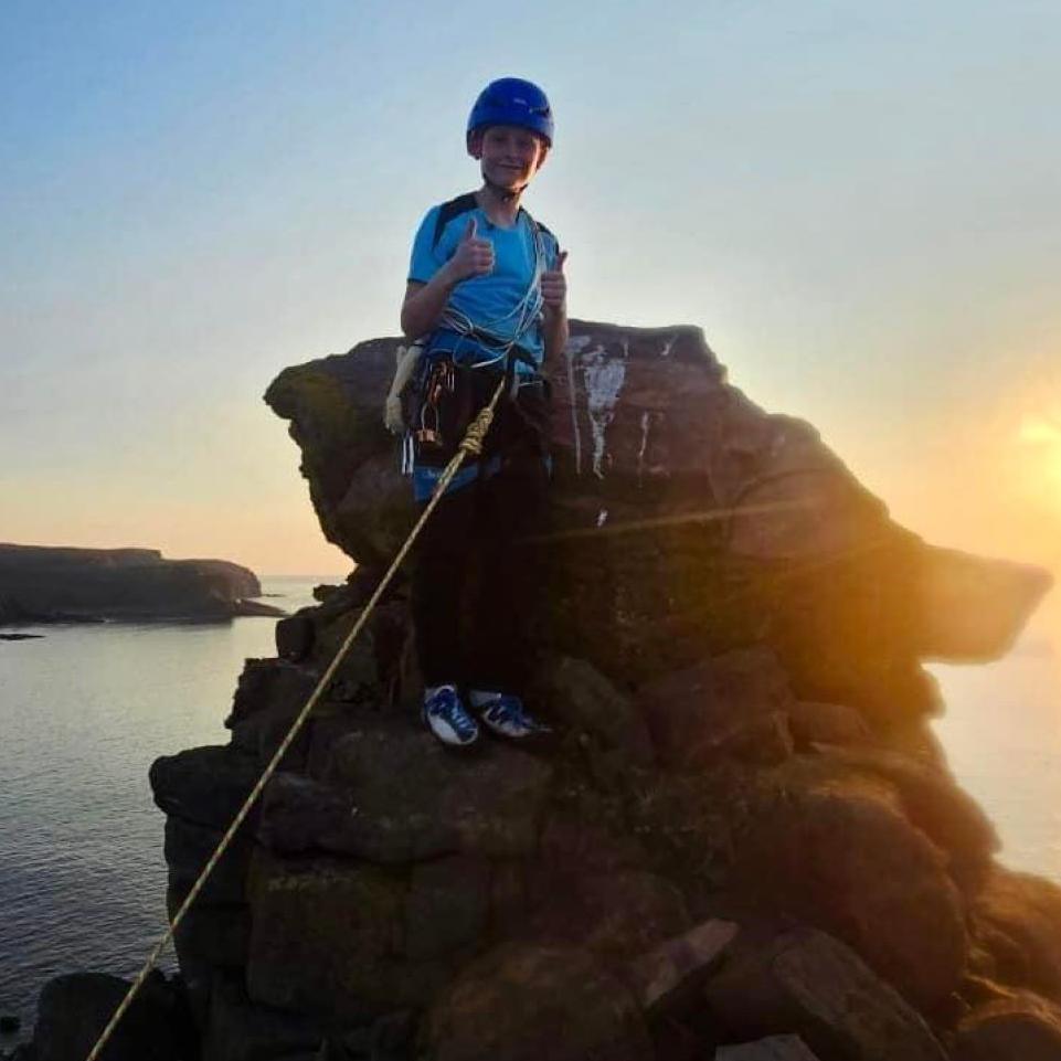 Aden Thurlow wearing climbing gear on top of the sea stack with the sun in the background